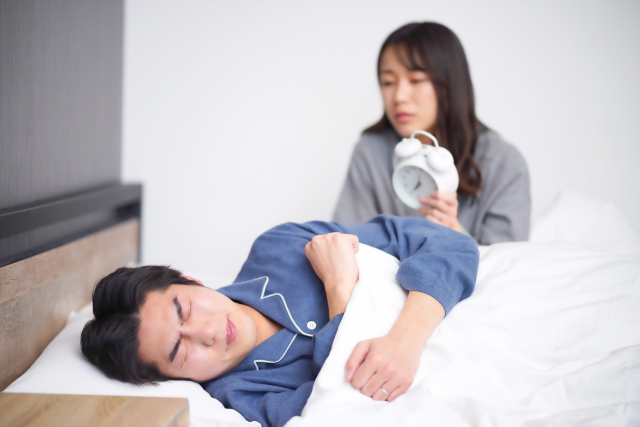 目覚まし時計を持って夫を起こそうとする妻